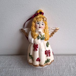 May include: A ceramic angel Christmas ornament. The angel has blonde hair, blue eyes, and gold-tipped wings. The white dress is decorated with red bows, green leaves, and red berries. A burgundy ribbon is attached for hanging.