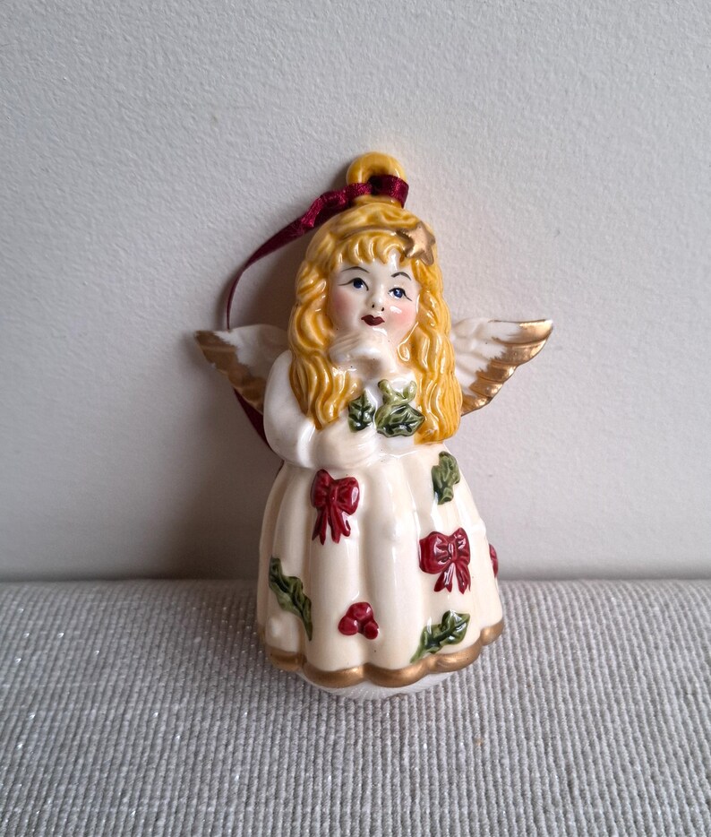 May include: A ceramic angel Christmas ornament. The angel has blonde hair, blue eyes, and gold-tipped wings. The white dress is decorated with red bows, green leaves, and red berries. A burgundy ribbon is attached for hanging.