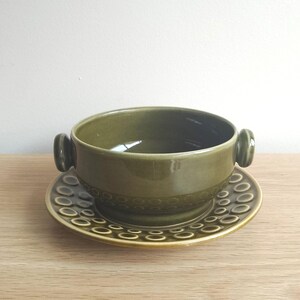 Puede incluir: Un tazón de sopa de cerámica verde con dos asas y un platillo a juego. El tazón y el platillo tienen un patrón texturizado de pequeños círculos.