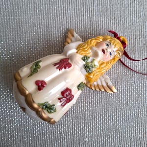 May include: A ceramic angel ornament with blonde hair, gold wings, and a cream-colored dress adorned with red bows and green leaves. The angel has a red ribbon for hanging and gold trim. A decorative holiday item.