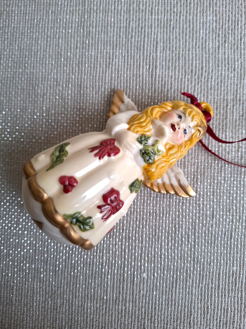 May include: A ceramic angel ornament with blonde hair, gold wings, and a cream-colored dress adorned with red bows and green leaves. The angel has a red ribbon for hanging and gold trim. A decorative holiday item.