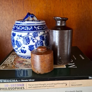 Könnte beinhalten: Ein Stillleben mit einem blau-weiß floral gemusterten Topf, einer braunen Steinzeugflasche und einem kleinen, runden, braunen Behälter. Die Objekte sind auf Büchern angeordnet.