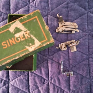 Könnte beinhalten: Eine offene grüne Schachtel mit dem Wort "SINGER" in orangefarbenen Buchstaben und mehrere silberne Nähmaschinenaufsätze auf einer violett gesteppten Oberfläche. Die Aufsätze umfassen Nähfüße und eine Schraube.