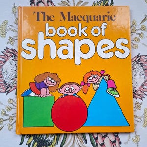 以下が含まれることがあります： 「The Macquarie book of shapes」というタイトルの子供向けの本で、鮮やかなオレンジ色の表紙です。表紙には、緑色の正方形、赤い球体、青い三角形など、幾何学的な形と交流する子供たちの漫画イラストが描かれています。