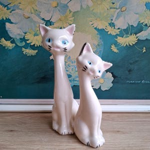 Pode incluir: Duas estatuetas de gato em cer&acirc;mica. Os gatos s&atilde;o brancos com olhos azuis, bigodes pretos e detalhes pretos nas orelhas e patas. Um gato tem um pesco&ccedil;o longo e esguio. O outro gato &eacute; mais curto e tem uma orelha com ponta preta. O fundo &eacute; uma pintura floral.