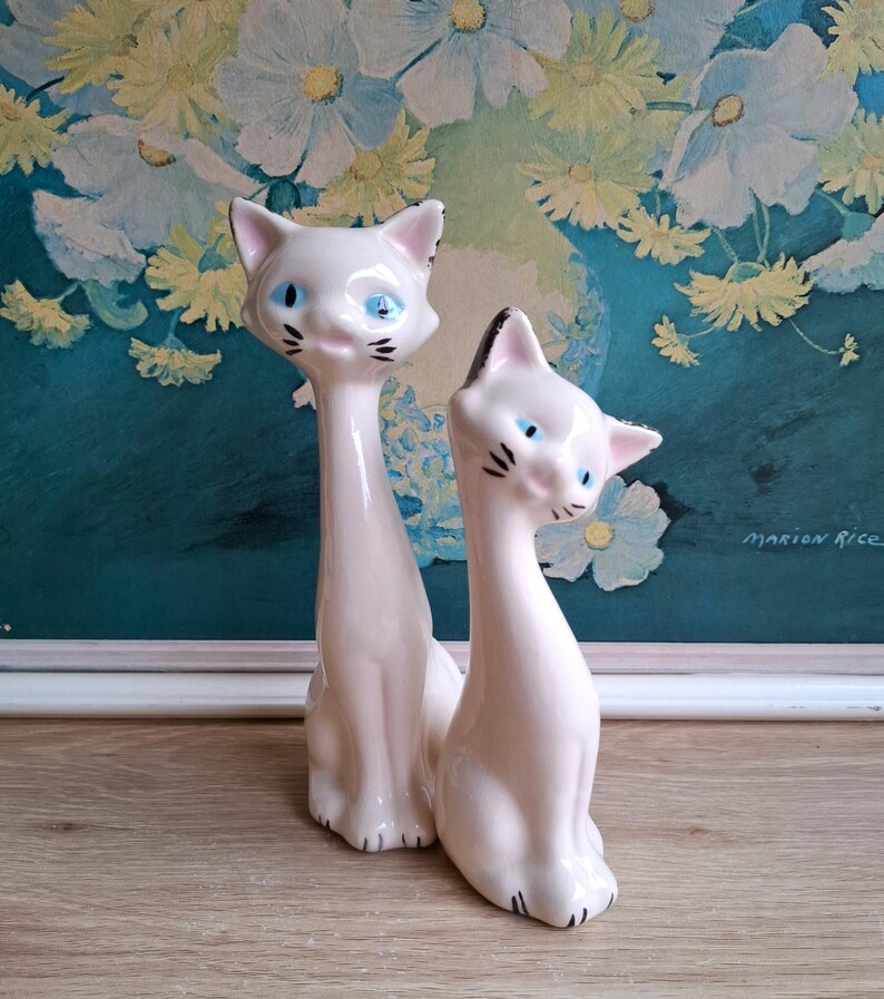 Pode incluir: Duas estatuetas de gato em cer&acirc;mica. Os gatos s&atilde;o brancos com olhos azuis, bigodes pretos e detalhes pretos nas orelhas e patas. Um gato tem um pesco&ccedil;o longo e esguio. O outro gato &eacute; mais curto e tem uma orelha com ponta preta. O fundo &eacute; uma pintura floral.