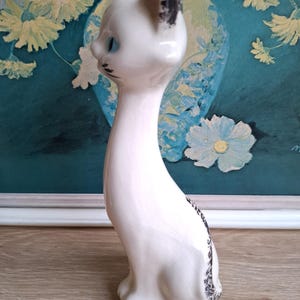 Pode incluir: Estatueta de gato em cer&acirc;mica branca com olhos azuis e detalhes em preto. O gato tem um pesco&ccedil;o longo e est&aacute; sentado em posi&ccedil;&atilde;o vertical. As orelhas e a cauda t&ecirc;m detalhes em preto. A estatueta est&aacute; em uma superf&iacute;cie de madeira.