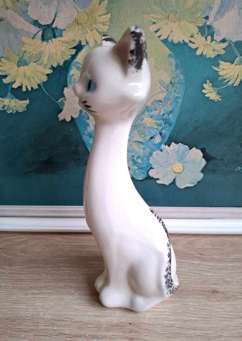 Pode incluir: Estatueta de gato em cer&acirc;mica branca com olhos azuis e detalhes em preto. O gato tem um pesco&ccedil;o longo e est&aacute; sentado em posi&ccedil;&atilde;o vertical. As orelhas e a cauda t&ecirc;m detalhes em preto. A estatueta est&aacute; em uma superf&iacute;cie de madeira.