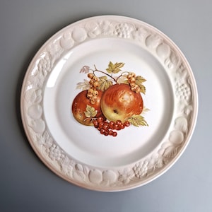 May include: White ceramic plate with a detailed fruit design. The plate features a central image of two apples and red currants, with a decorative embossed border of fruits and leaves. A classic piece for serving.