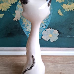 Pode incluir: Estatueta de gato em cer&acirc;mica branca com marcas pretas nas orelhas e um redemoinho preto nas costas. O gato tem um pesco&ccedil;o longo e esguio e um corpo arredondado. A estatueta &eacute; colocada em frente a uma pintura floral.