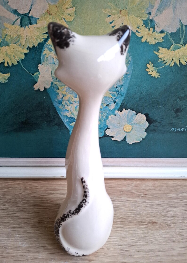 Pode incluir: Estatueta de gato em cer&acirc;mica branca com marcas pretas nas orelhas e um redemoinho preto nas costas. O gato tem um pesco&ccedil;o longo e esguio e um corpo arredondado. A estatueta &eacute; colocada em frente a uma pintura floral.