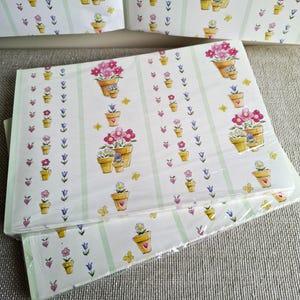 May include: Sheets of floral-themed wrapping paper. The paper features a repeating pattern of potted flowers in shades of pink, yellow, and purple, along with butterflies and hearts. The background is white with vertical green stripes.