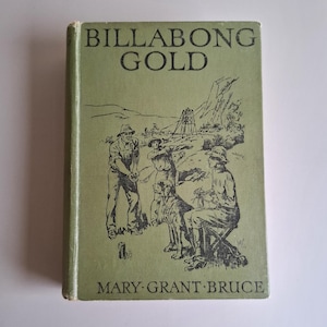 Op de afbeelding: Een vintage boek getiteld "BILLABONG GOLD" met een groene kaft. De kaft heeft een zwart-wit illustratie van verschillende mensen, een hond en een mijnstructuur. De naam van de auteur, MARY GRANT BRUCE, staat onderaan.