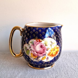 May include: A vintage blue ceramic pitcher with a gold handle and trim. The pitcher features a floral design with pink and yellow roses on a white cloud-shaped background. The background is navy blue with gold swirl details.