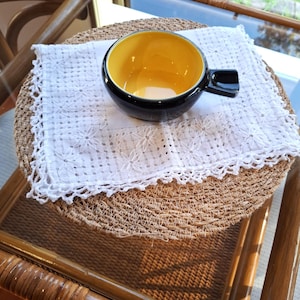 Pode incluir: Uma pequena tigela redonda de cerâmica com exterior preto brilhante e interior amarelo vivo, apresentando uma única alça curta e quadrada na lateral, apoiada sobre um guardanapo de tecido branco quadrado com bordado floral complexo e bordas de renda.