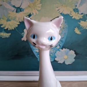 Pode incluir: Estatueta de gato em cer&acirc;mica branca com pesco&ccedil;o comprido e olhos azuis. O gato tem marcas pretas nas orelhas e no rosto. A estatueta &eacute; colocada em um fundo de pintura floral.