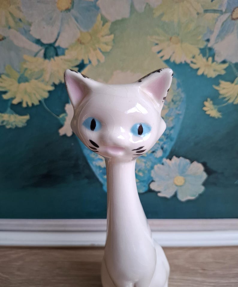 Pode incluir: Estatueta de gato em cer&acirc;mica branca com pesco&ccedil;o comprido e olhos azuis. O gato tem marcas pretas nas orelhas e no rosto. A estatueta &eacute; colocada em um fundo de pintura floral.