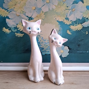 Pode incluir: Duas figuras de gato de cer&acirc;mica vintage. Os gatos s&atilde;o brancos com detalhes pretos nas orelhas, nariz e bigodes. T&ecirc;m olhos azuis e est&atilde;o sentados. O fundo apresenta uma pintura floral com flores brancas e amarelas.