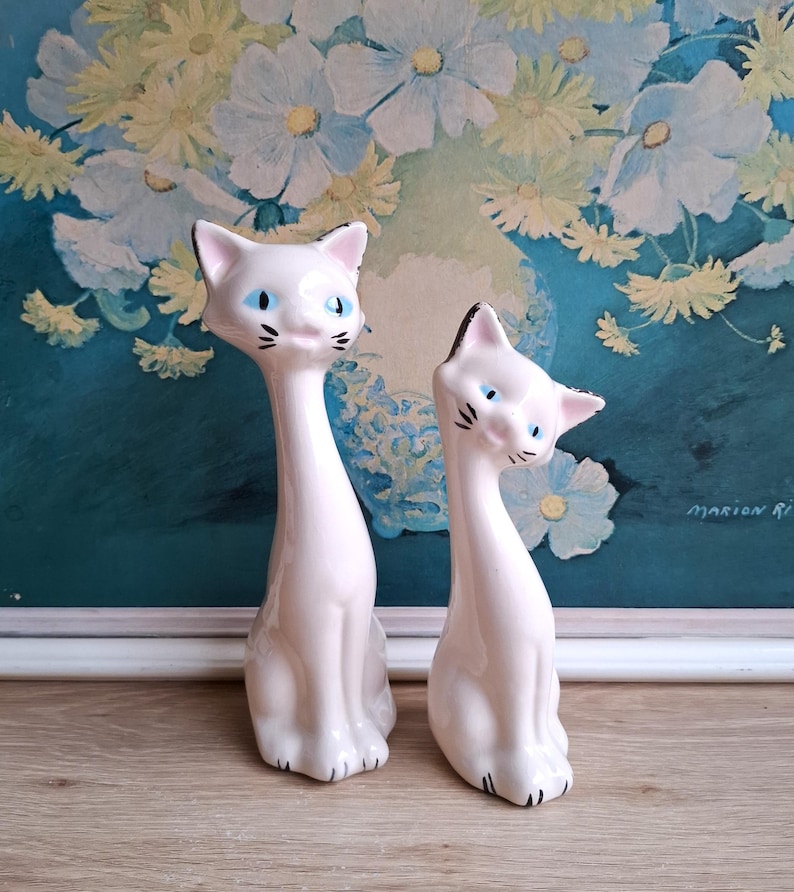 Pode incluir: Duas figuras de gato de cer&acirc;mica vintage. Os gatos s&atilde;o brancos com detalhes pretos nas orelhas, nariz e bigodes. T&ecirc;m olhos azuis e est&atilde;o sentados. O fundo apresenta uma pintura floral com flores brancas e amarelas.