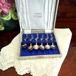 May include: A vintage set of silver spoons presented in a blue and gold box. The box is lined with blue fabric and white satin. The spoons have decorative floral handles. The box rests on a white lace doily.