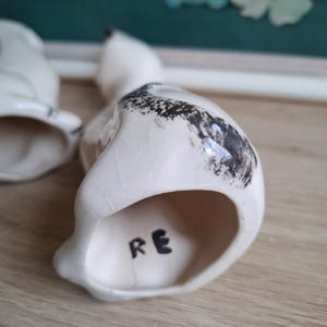 Pode incluir: Estatueta de p&aacute;ssaro em cer&acirc;mica branca com detalhes salpicados em preto nas costas. As letras "RE" s&atilde;o pintadas &agrave; m&atilde;o em preto na parte inferior. A estatueta est&aacute; sobre uma superf&iacute;cie de madeira.