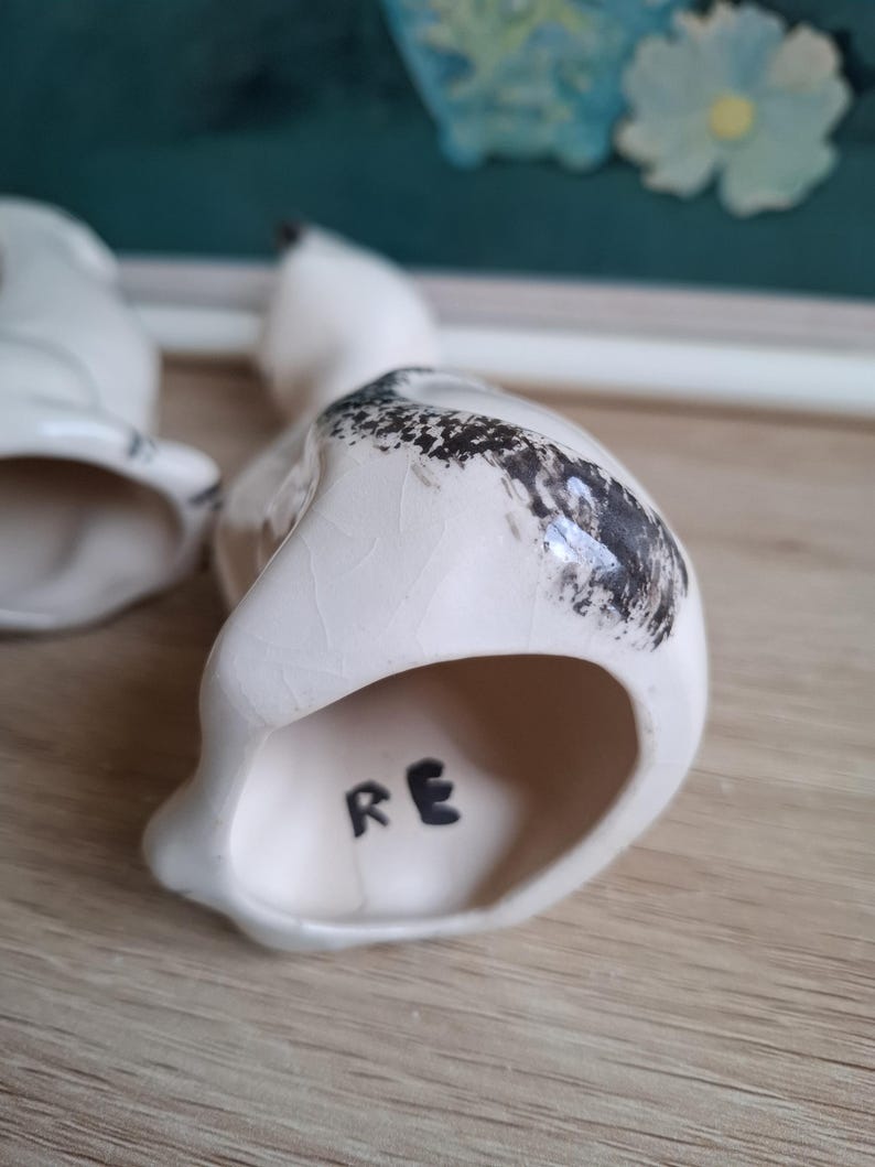 Pode incluir: Estatueta de p&aacute;ssaro em cer&acirc;mica branca com detalhes salpicados em preto nas costas. As letras "RE" s&atilde;o pintadas &agrave; m&atilde;o em preto na parte inferior. A estatueta est&aacute; sobre uma superf&iacute;cie de madeira.