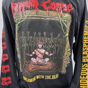 Puede incluir: Camiseta negra de manga larga con un diseño gráfico que presenta el texto "VIOLENT CORPSE" en letras rojas goteantes. El diseño muestra una figura meditando frente a esqueletos, con las palabras "DREAMING WITH THE DEAD". La manga derecha tiene el texto "HIDEOUS BLASPHEMIES".