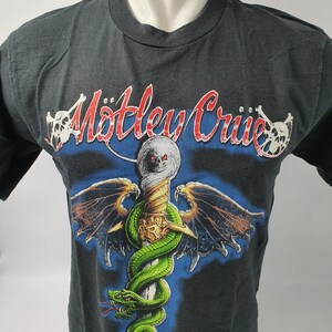 以下が含まれることがあります： 黒いTシャツに、翼のある剣に巻き付いたヘビのグラフィックプリントが施されています。グラフィックの上には「Motley Crue」という文字が赤と白でプリントされています。