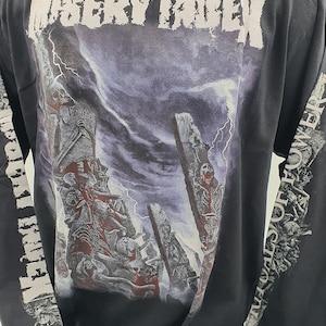 May include: Black long-sleeve t-shirt with a graphic print of a skeletal figure in front of a stormy sky. The text "Misery Index" is printed across the top of the shirt. The text "Veritas" is printed on the graphic. The text "The Walls Of Power" is printed on the left sleeve.