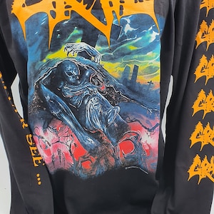 May include: Black long-sleeve shirt featuring the band name "GRAVE" in orange, stylized lettering across the chest. The shirt's graphic depicts a dark, detailed illustration with a skeletal figure and a graveyard scene. The sleeves have an orange design.