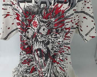 May include: White t-shirt with a graphic print of a bloody, screaming face with chains and the word "TESTAMENT" in large, gray letters. The background is white with red blood splatters.