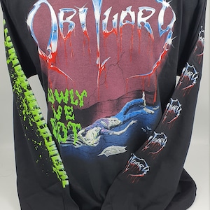 Könnte beinhalten: Schwarzes Langarm-T-Shirt mit einem Grafikdruck der Band Obituary. Die Grafik zeigt den Bandnamen in einer metallischen Schriftart mit tropfendem Blut und eine Szene eines am Boden liegenden Körpers. Die Ärmel haben eine sich wiederholende Grafik des Bandnamens in grüner Schrift.