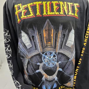 Pode incluir: Camiseta preta de manga comprida com a palavra "PATIENCE" em letras de bloco amarelas e vermelhas. A camisa apresenta um gráfico detalhado de uma estrutura gótica com uma porta central e um design circular complexo. As mangas têm gráficos brancos.