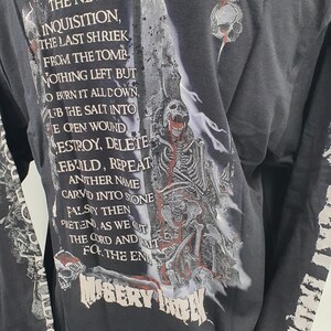 May include: Black long-sleeve t-shirt with a graphic print of a skeleton holding a skull. The text "WELCOME ALL TO THE NEW INQUISITION, THE LAST SHRIEK FROM THE TOMB NOTHING LEFT BUT O BURN IT ALL DOWN, UB THE SALT INTO THE OPEN WOUND DESTROY, DELETE REBUILD, REPEAT ANOTHER NAME CARVED INTO STONE FALSEY THEN PRETEND, AS WE CUT THE CORD AND WAIT FOR THE END MISERY INDEX" is printed on the back of the shirt.