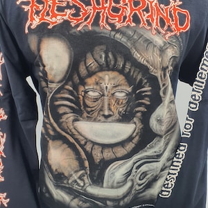 May include: Black long-sleeve t-shirt with a graphic print of a creature with a large head and a detailed face. The text "FLESHGRIND" is printed in red on the front of the shirt. The text "Destined for torment" is printed on the right sleeve.
