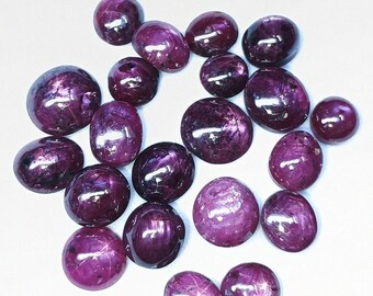 May include: A collection of polished, oval-shaped gemstones in varying sizes. The gems are a deep, rich purple color with a glossy finish, scattered against a white background. The stones appear to be cabochons.