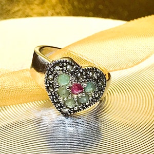 May include: A silver heart-shaped ring with a red gemstone surrounded by green gemstones and black marcasite stones.