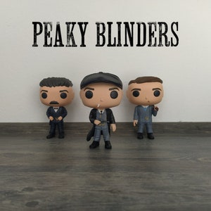 以下が含まれることがあります： ピーキー・ブラインダーズのファンコポップフィギュア3体。フィギュアはトミー・シェルビー、アーサー・シェルビー、ジョン・シェルビーです。フィギュアは特徴的なスーツと帽子を着用しています。フィギュアは木製の表面に立っています。背景は白い壁で、「ピーキー・ブラインダーズ」という文字が黒で書かれています。