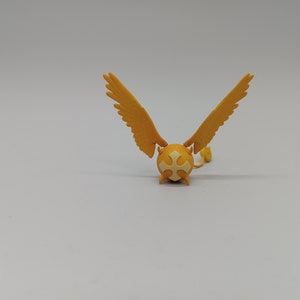 Pode incluir: Uma figurinha amarela com asas e uma cruz branca nas costas. A figurinha é feita de plástico e tem uma pequena cauda.