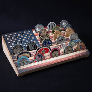 May include: A wooden coin display case with an American flag design. The case has slots for 20 coins and features a variety of challenge coins with different designs and colors.