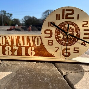 May include: A wooden clock with a brown and white design. The clock face features the text "POLICIA DE PUERTO RICO EN LA FLORIDA" and "PASADIA FAMILIAR". The clock is mounted on a wooden base with the text "MONTALVO 18716".