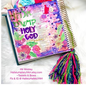 May include: A colorful, decorated journal with the words "HOLY GOD" and Hebrew text. The journal features floral illustrations, a gold spiral binding, and a tassel. The cover is adorned with various fabric embellishments.