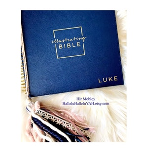 Pode incluir: Um diário de couro azul marinho com letras douradas que dizem "Illustrating Bible" e "Luke". O diário tem uma encadernação em espiral dourada e um tassel com detalhes em rosa, branco e azul.