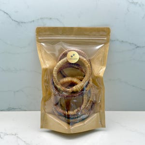 May include: A clear plastic bag filled with several dried dog treats. The treats are circular, light brown, and appear to be made of natural ingredients. A small sticker with the words "noble paws" is on the bag.