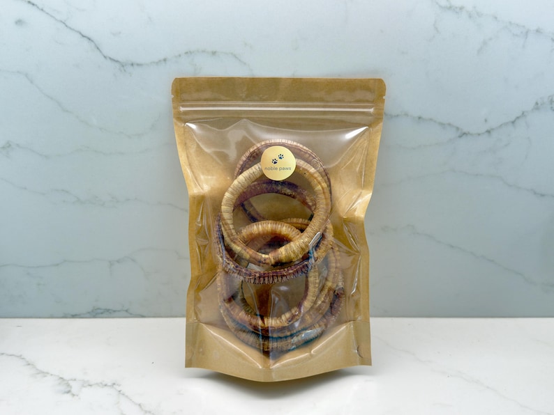 May include: A clear plastic bag filled with several dried dog treats. The treats are circular, light brown, and appear to be made of natural ingredients. A small sticker with the words "noble paws" is on the bag.