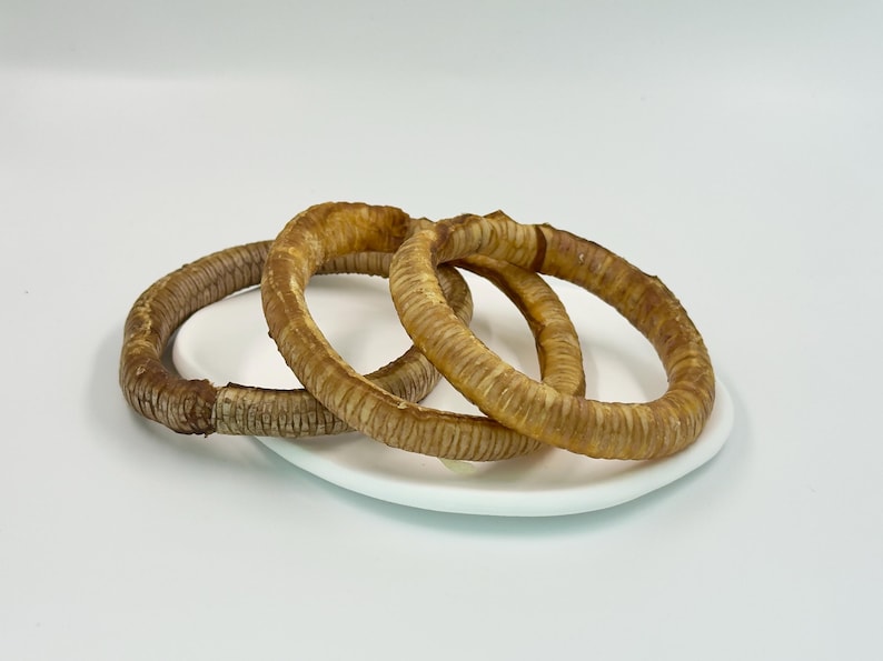May include: Three dried, ring-shaped dog treats with a textured surface, in shades of brown and tan, are arranged on a white plate. The treats are positioned against a plain white background, highlighting their shape and color.