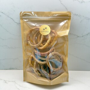 May include: A clear plastic bag filled with multiple tan-colored, circular dog chews. The bag has a gold-colored label with the words "noble paws" and paw prints. The background is a white and gray marble pattern.