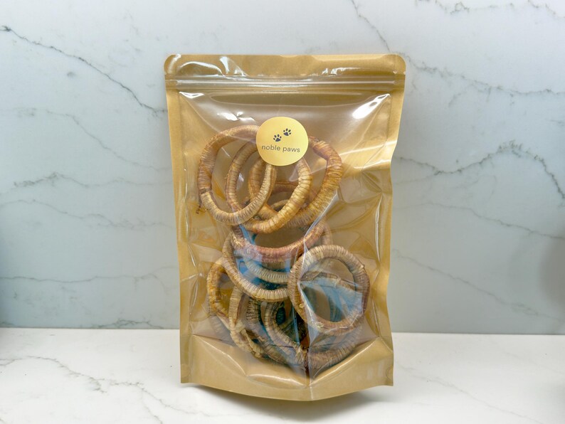 May include: A clear plastic bag filled with multiple tan-colored, circular dog chews. The bag has a gold-colored label with the words "noble paws" and paw prints. The background is a white and gray marble pattern.