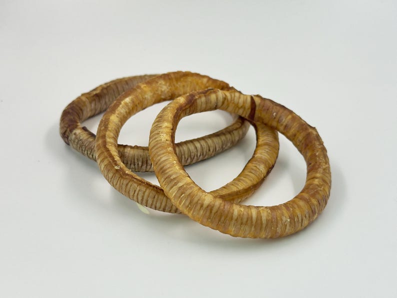 May include: Three circular, dried dog treats in shades of brown and tan are arranged on a white surface. The treats have a textured, ridged appearance, and are likely made from natural ingredients.