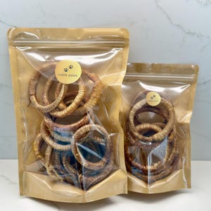 May include: Two clear plastic bags filled with dog treats. The bags are brown and sealed with a zipper. The treats are ring-shaped and brown. Each bag has a gold sticker with the words "noble paws" and paw prints.